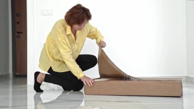 Parlak sarı bluzlu bir kadın heyecan verici bir şekilde yeni mobilya kutusunu açıyor. Tüm parçaları kontrol eder ve montaj için hazırlanır. Yeni yaşam alanı için her detayın düzenli olduğundan emin olur..