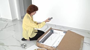 Bir kadın yeni aldığı mobilyaları parlak bir odada monte etmeye hazırlanıyor. Kutuyu dikkatlice açıyor, parçaları düzenliyor ve kuruluma başlamadan önce gerekli tüm aksesuarları kontrol ediyor..