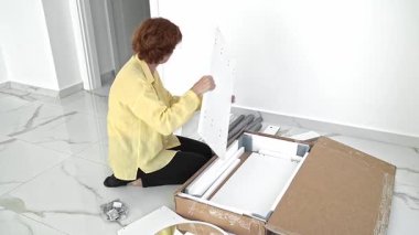 Bir kadın heyecanlı bir şekilde yeni mobilya parçalarıyla dolu bir kutuyu açar, şık dairesinde montaj işlemine başlamadan önce tüm parçaları ve aksesuarları dikkatle kontrol eder, yeni bir eve taşınmaya hazırlanır..