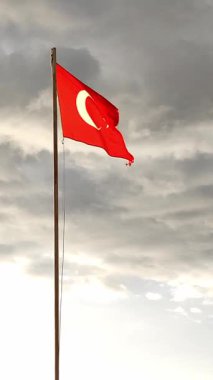 Türkiye 'nin canlı bir kırmızı bayrağı, parlak beyaz bir yıldız ve hilal şeklinde görünen bir bayrak direğinin üzerinde görkemli bir şekilde açılır. Arkaplanda çarpıcı bayrakları yükselten dramatik bulutlar yer alıyor.
