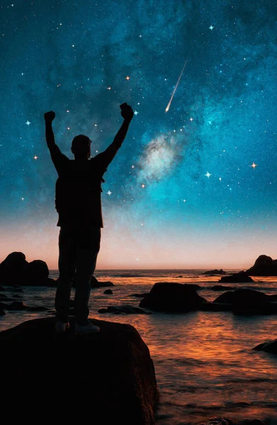 silhouette of a person on a rock hands up at night with milky way and stars on the background over the ocean