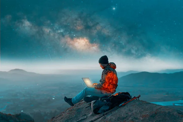 stock image silhouette of a person sitting on top of a mountain working on his laptop at night over the valley