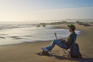 Dijital göçebe olgun bir adam plajda oturmuş laptopuyla rahatça çalışıyor.