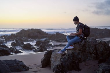 Olgun erkek dijital göçebe gün batımında plajda dizüstü bilgisayarıyla çalışıyor.