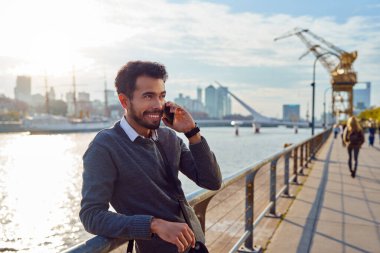 Latin Amerikalı genç, yetişkin bir işadamı cep telefonuyla konuşuyor. Arka planda bir nehirle sokakta gülümsüyor.