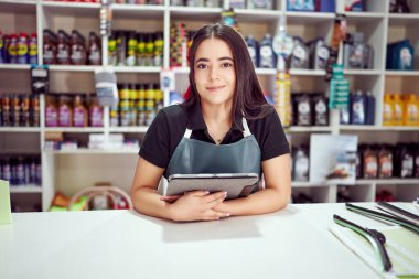 Latin Amerikalı kadın tezgahın önünde duruyor ve elinde küçük otomobil parçaları satan dijital bir tablet var.