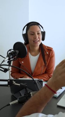 İki radyo sunucusu bir stüdyoda canlı yayın sırasında sohbet ediyorlar. Masada oturan kulaklık takan erkek ve kadın kayıt için dizüstü bilgisayar ve tablet bilgisayar kullanıyor. 