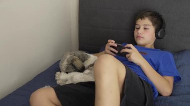 Kulaklık takan çocuk, yanında köpeği yatarken yatakta video oyunu oynamaktan hoşlanıyor.