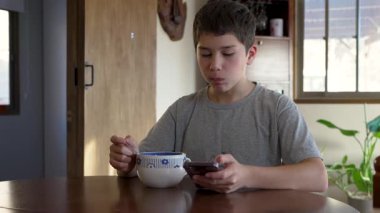 Çocuk mutfak masasındaki kaseden kahvaltı gevreği yiyor ve akıllı telefon kullanıyor. 
