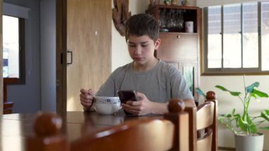 Çocuk mutfak masasındaki kaseden kahvaltı gevreği yiyor ve akıllı telefon kullanıyor. 