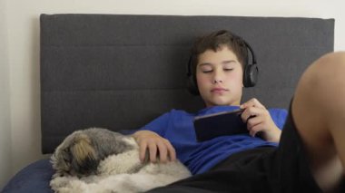 Kulaklık takan çocuk yatakta video oyunu oynamaktan, köpeğini okşamaktan hoşlanıyor.
