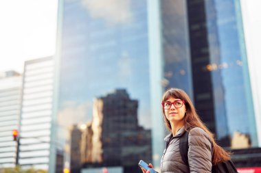 Elinde cep telefonuyla şehirde binalarla dolaşan güzel, genç, beyaz bir kadının portresi.
