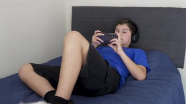 Çocuk kulaklık takmış, akıllı telefonuyla video oyunu oynuyor ve yatağında yatıyor.