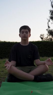 Rahat çocuk gün batımında dışarıda meditasyon yapıyor, yoga minderinde çimlerin üzerinde oturuyor.