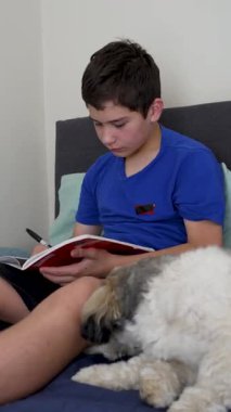 Kitaplarda yazan konsantre ergen çocuk köpeğiyle yatakta oturuyor.