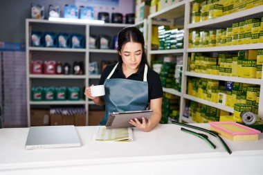 Genç Latin Amerikalı kadın küçük bir oto yedek parça dükkanında tablet tutuyor ve kahve içiyor.