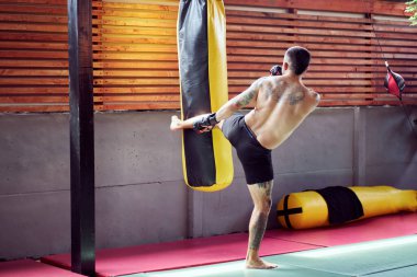 Erkek kickboksör spor salonunda döner tekme antrenmanı yapıyor, antrenman sırasında kum torbasına vuruyor.