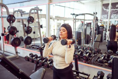 Spor salonunda dambıllarla egzersiz yapan Latin bir genç kadın.