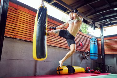 Kaslı kickboksör, dinamik bir spor salonunda gücünü ve tekniğini sergileyerek ağır bir torbaya güçlü bir tekme atıyor.