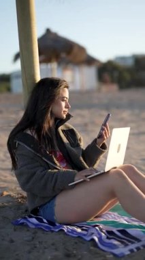 Güzel Latin bir kadın kumlu bir sahilde dizüstü bilgisayar ve akıllı telefon kullanarak oturuyor.