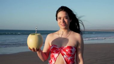 Gün batımında plajda kavun meyvesi içinde kavun içen mutlu genç kadın, tatil ve yaz konsepti.