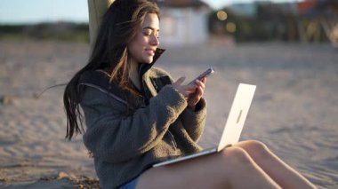 Güzel Latin bir kadın kumlu bir sahilde dizüstü bilgisayar ve akıllı telefon kullanarak oturuyor.