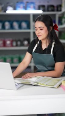 Latin Amerikalı genç bir kadın dizüstü bilgisayar kullanıyor ve küçük bir oto yedek parça şirketinde ürün listesine bakıyor.