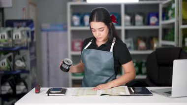 Latin Amerikalı genç bir kadın konserve kutusu tutuyor ve küçük bir oto yedek parça şirketindeki ürün listesine bakıyor.