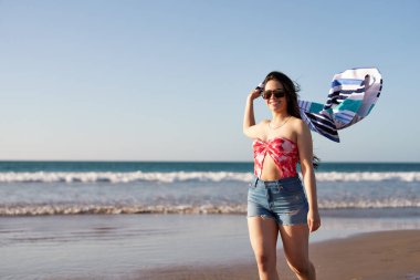 Kaygısız genç bir kadın kumsalda elinde bir elbiseyle yürüyor yaz tatilinin tadını çıkarıyor.