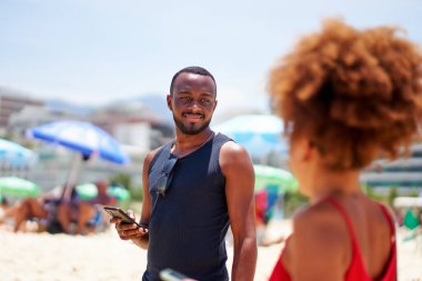 Genç siyah Brezilyalı bir adam elinde bir cep telefonu tutuyor ve sahildeki bir kadına bakıyor.