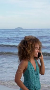 Kıvırcık saçlı genç Afrikalı kadın deniz kıyısında cep telefonuyla konuşuyor.