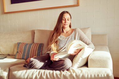 Güzel beyaz kadın gün batımında evde kanepede kitap okuyor. Sağlık ve kendine bakım kavramı.