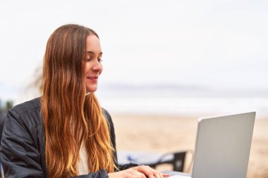Sahilde dizüstü bilgisayar kullanarak oturan beyaz güzel bir kadının portresi.