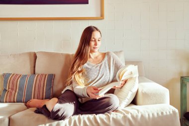 Genç, beyaz bir kadın evdeki kanepede kitap okuyor. Sağlık ve kendine bakım kavramı.