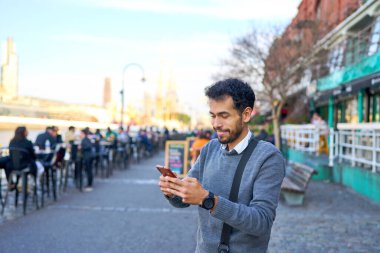 Latin Amerikalı genç işadamı cep telefonuna bakıyor ve şehirde gülümsüyor.