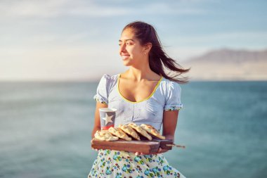 Tipik huasa kostümü giymiş Latin bir kadının portresi. Elinde çam böreği ve üzerinde Şili bayrağı olan geri dönüşümlü bir bardak var.