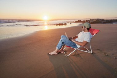 Dijital göçebe sahilde oturuyor ve güzel gün batımında dizüstü bilgisayarıyla çalışıyor.