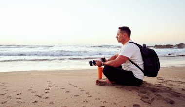 Fotoğrafçı elinde kamerayla kumsalda dinleniyor. Gün batımında huzurlu okyanus manzarasının tadını çıkarıyor.