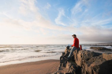 Olgun Latin adam sahildeki kayada dizüstü bilgisayarıyla oturuyor ve günbatımının tadını çıkarıyor.