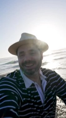 Hasır şapkalı mutlu adam sahilde günbatımının tadını çıkarıyor, denizi gösteriyor.