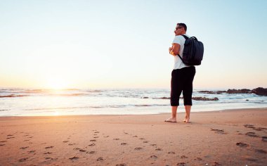 Sırt çantalı erkek turist kumlu sahilde güzel bir altın gündoğumunun tadını çıkarıyor. Seyahat ve maceralarla dolu yeni bir güne başlıyor.