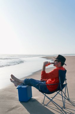 Kumsalda oturmuş bira içen olgun Latin bir adam. 