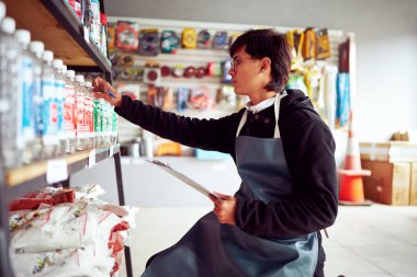 Konsantre olmuş erkek tezgahtar nalbur dükkanında stok yapıyor ve pano ile stokları kontrol ediyor.