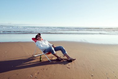Olgun bir adam plaj sandalyesinde uzanmış, gözleri sahilde kapalıyken rahatlamış.