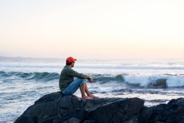 Genç adam gün batımında sahilde kayada tek başına oturuyor.