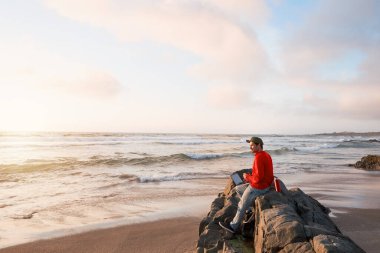 Kumsaldaki kayalarda oturan olgun Latin bir adam elinde dizüstü bilgisayar ve açık havada bir fincan kahveyle. 