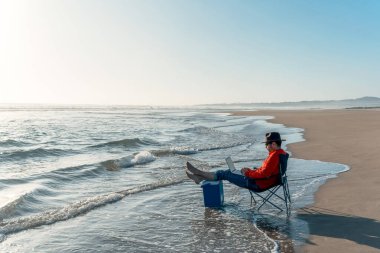 Dijital göçebe dizüstü bilgisayarla oturuyor ve deniz kıyısında çalışıyor