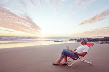 Genç adam tropikal bir plajda dizüstü bilgisayarla çalışıyor. Serbest ve iş konseptinde.