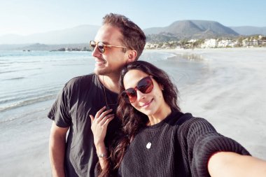 Mutlu Latin heteroseksüel çift sahilde selfie çekiyor, kumsalın tadını çıkarıyor.