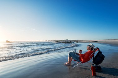 Kumsalda dizüstü bilgisayarıyla oturmuş kahve içen olgun Latin bir adam.